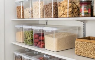 Transparent kitchen storage containers neatly arranged on shelves, filled with grains, pulses, snacks, and dry fruits. Plastic storage boxes