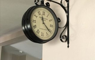 A vintage-style double-sided wall clock with Roman numerals, mounted on a decorative wrought iron bracket. Sitting room wall clock